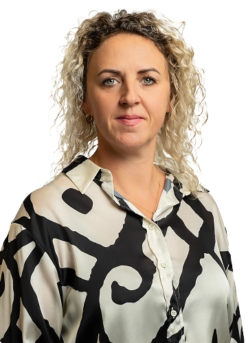 Kobieta o kręconych blond włosach pozuje do zdjęcia w koszuli z czarno-białym wzorem.