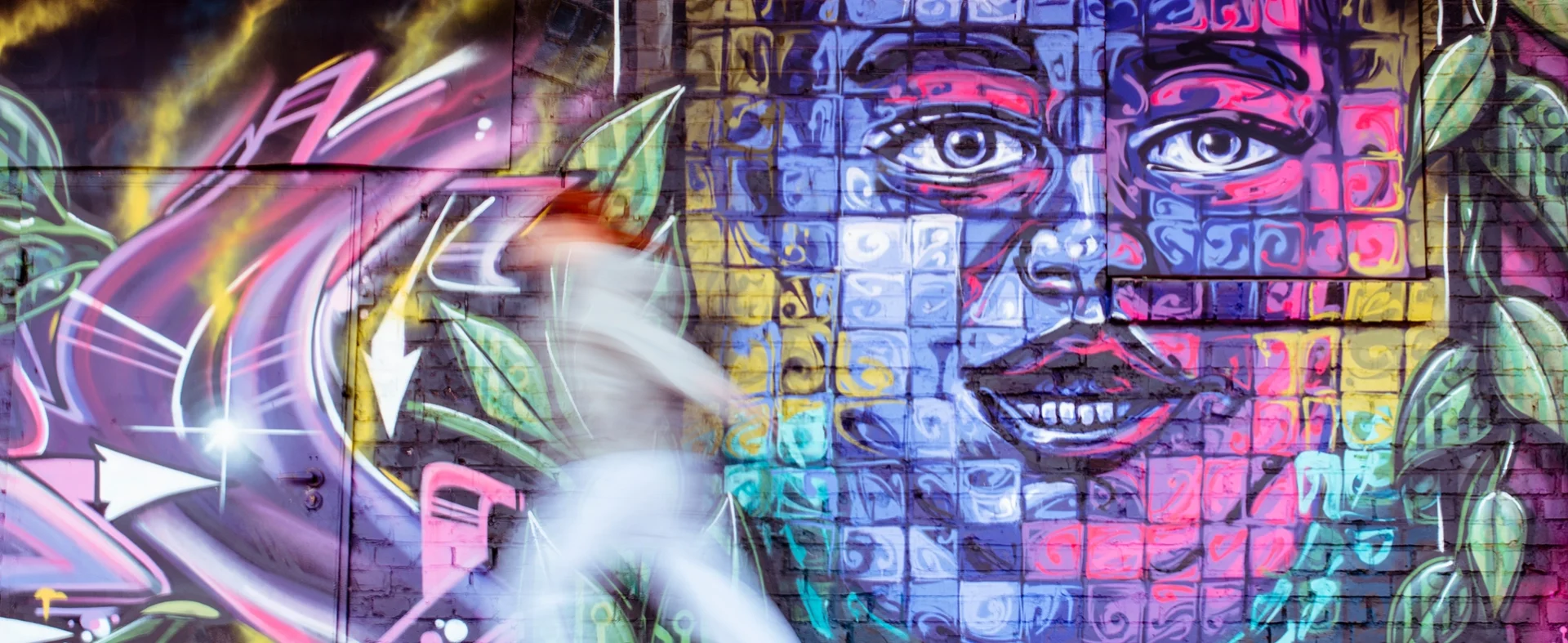 Kolorowy mural z twarzą na ścianie, obok której rozmazana sylwetka osoby idzie.